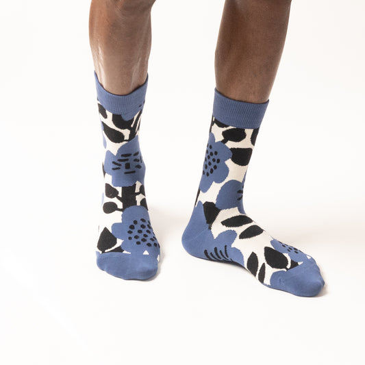 Front view of a person's lower legs in periwinkle blue crew socks with a large black and cream floral and leaf print, one hand resting on the knee, on a white background.