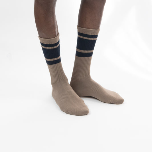 A pair of beige crew socks with two navy blue horizontal stripes near the ribbed cuffs, shown on a model's feet against a white studio background.