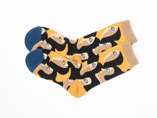A pair of crew socks with a bold graphic pattern of stylized faces with closed eyes on a black background, accented with mustard yellow shapes, beige cuffs and heels, and solid blue toes, displayed on a clean white background.