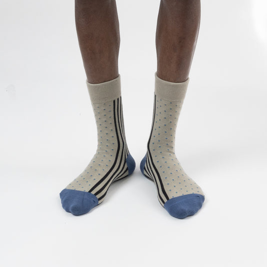 A pair of crew socks worn on a model's legs, featuring a light beige base with thin black vertical stripes, small blue polka dots, and solid blue toes and heels, photographed against a seamless white background.
