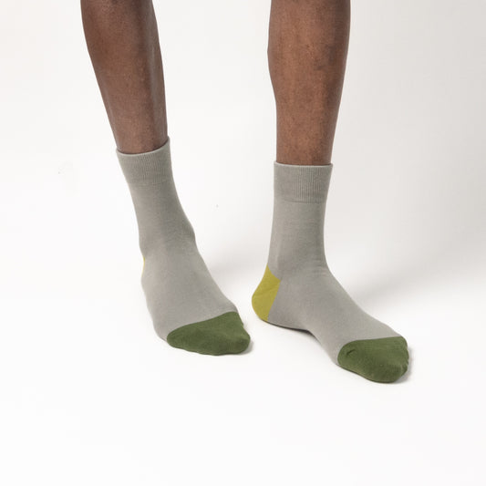 A close-up front view of a person's lower legs and feet wearing crew-length socks. The socks are primarily a light gray color, featuring a solid dark green patch on the toes and a small lime green accent visible near the heel. The background is solid white.