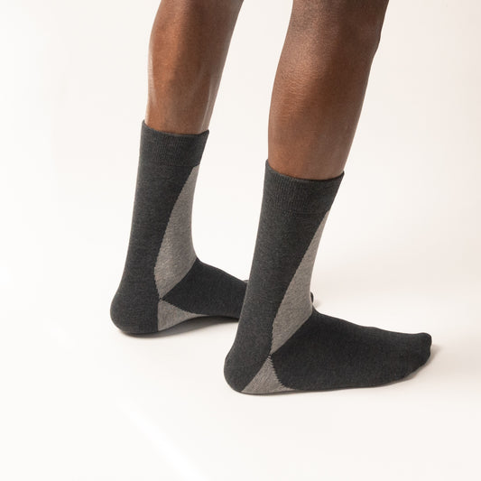 Model standing in charcoal grey crew socks with a light grey geometric color-block panel on the side and heel, shown from a rear three-quarter angle against a white background.