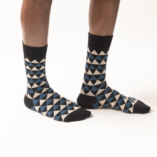 Model standing in black crew socks with a geometric triangle pattern in steel blue and cream, black cuffs, heels, and toes, shown from a three-quarter angle against a white background.