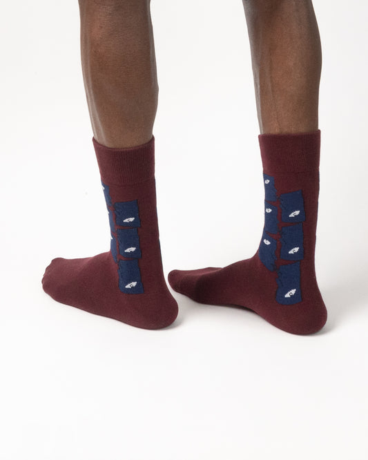A pair of crew socks worn on a person's feet, shown from the back against a clean white studio background. The socks are burgundy with a navy blue heel panel featuring small eye motifs.