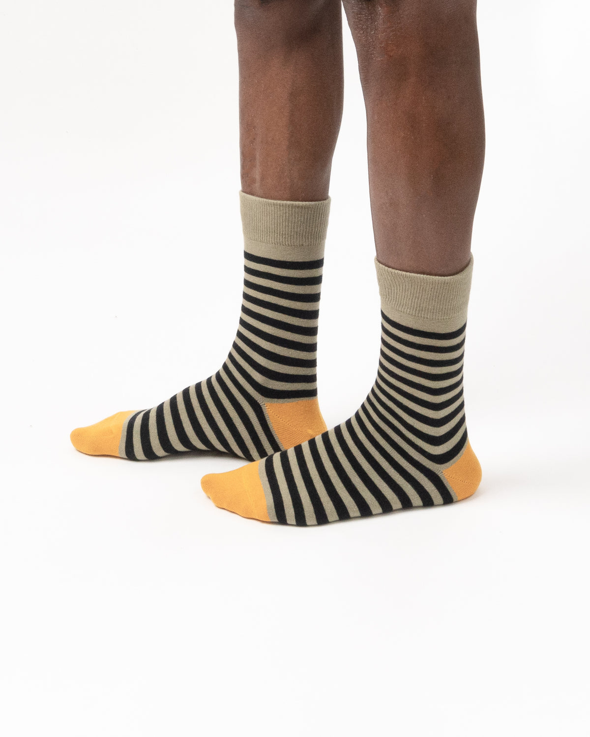 A pair of crew socks with horizontal black and beige stripes, featuring solid yellow toes and heels, shown on a model's feet against a white background.