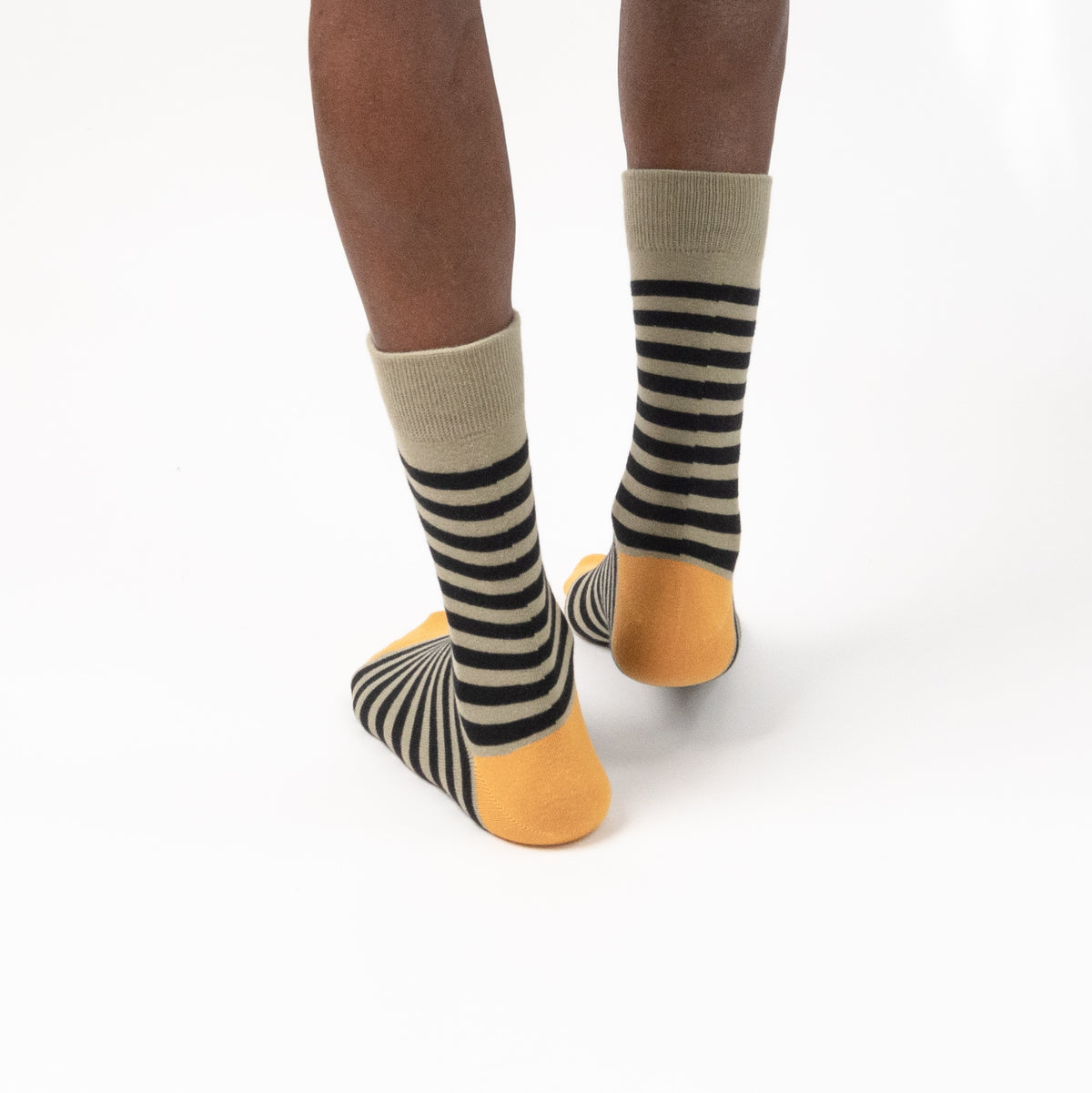 A person's feet and lower legs wearing striped crew socks with a beige cuff, black and beige diagonal stripes, and a mustard yellow toe and sole, shown against a plain white background.