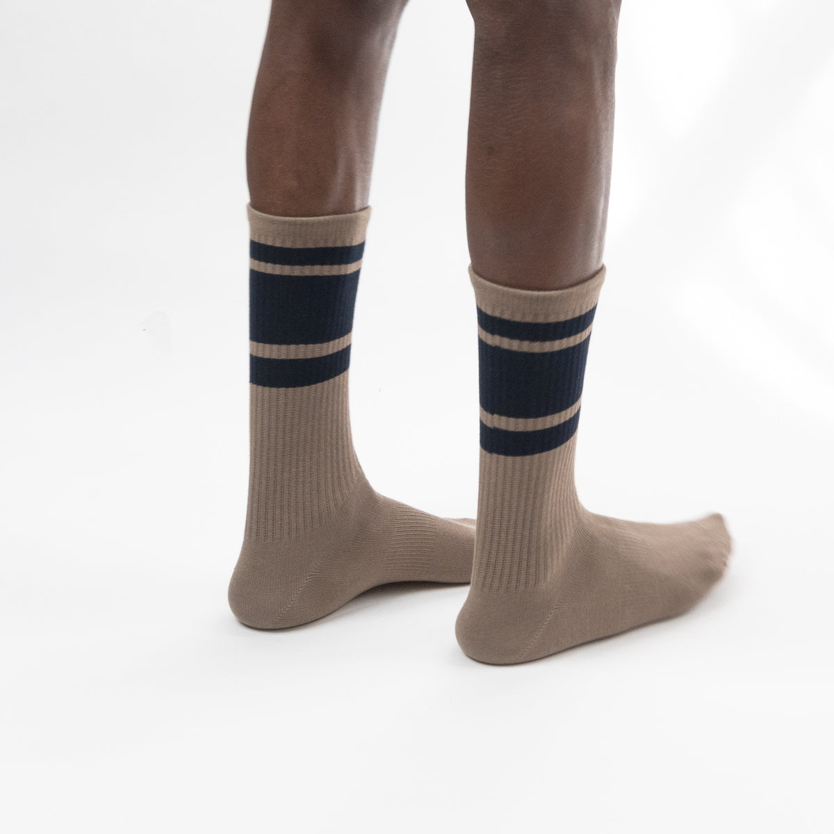 A pair of beige crew socks with navy blue stripes near the top, worn on a person's legs against a plain white studio background.