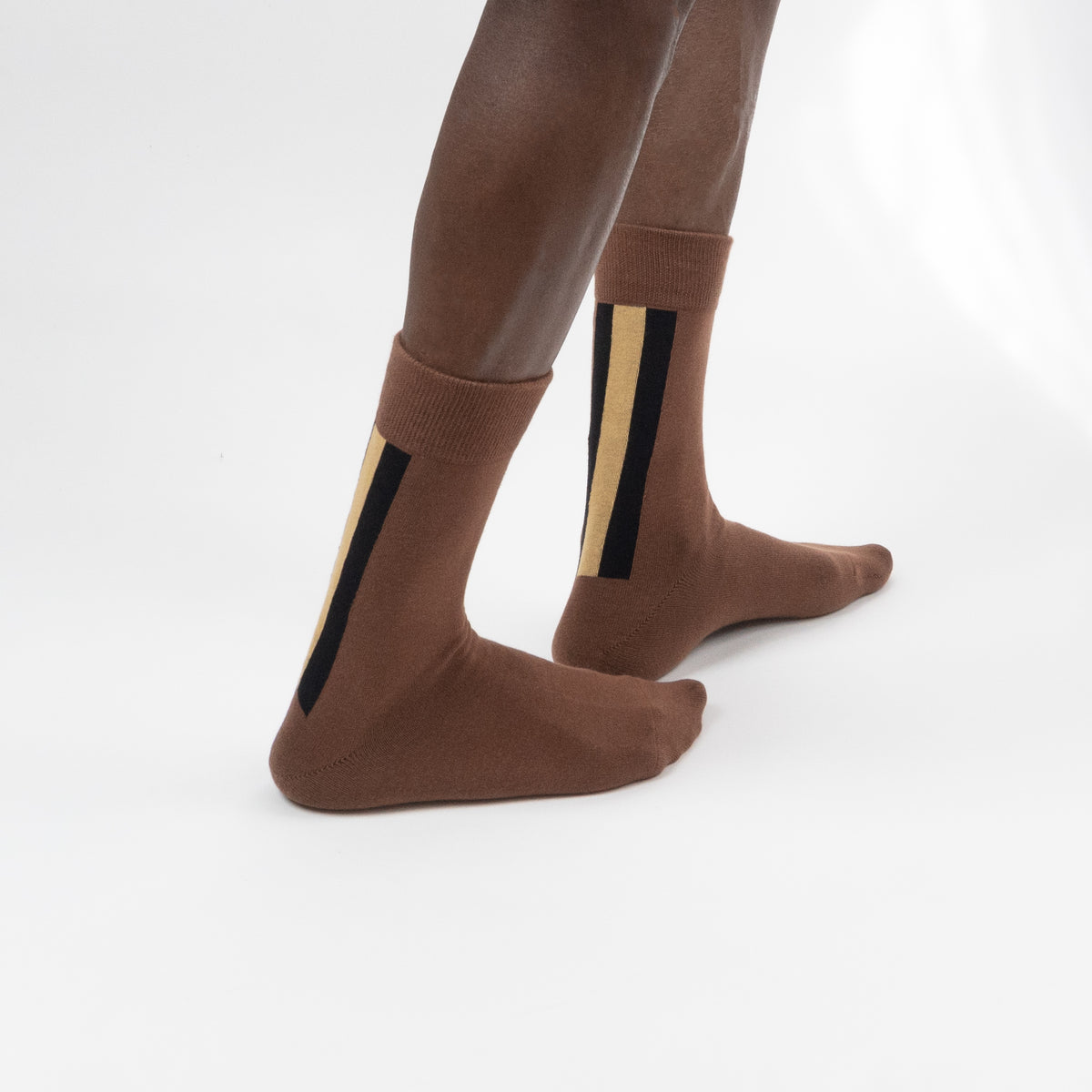 Three-quarter back view of a model wearing mid-calf brown crew socks featuring thick vertical black and tan accent stripes on the side of the ankle, standing against a white background.
