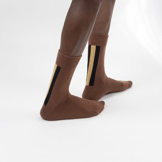 Three-quarter back view of a model wearing mid-calf brown crew socks featuring thick vertical black and tan accent stripes on the side of the ankle, standing against a white background.