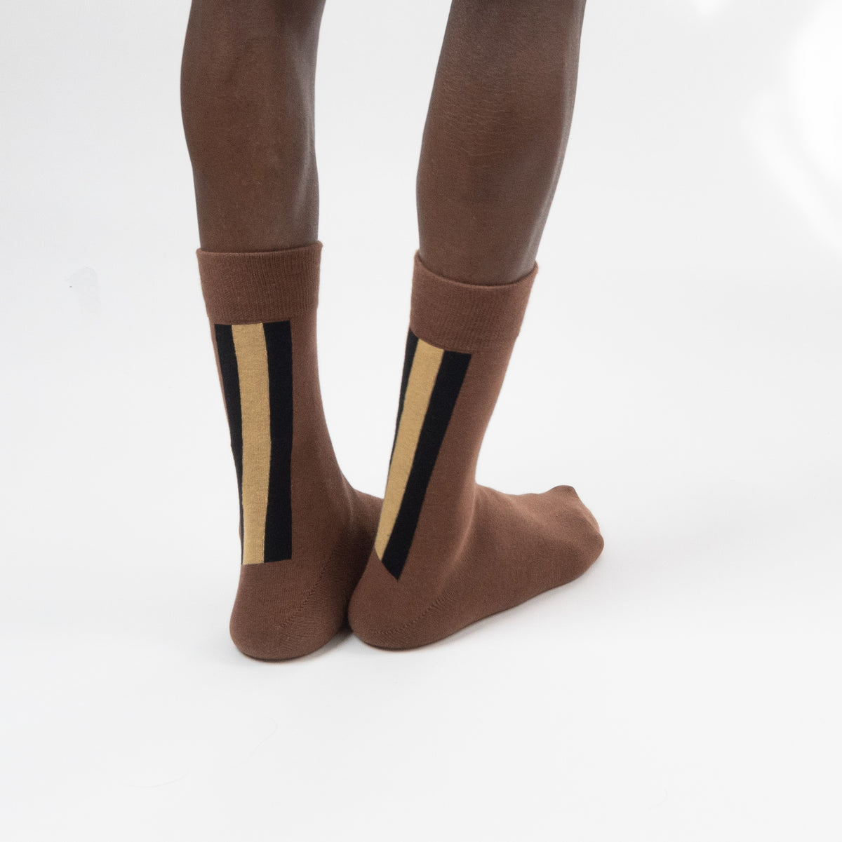 A pair of brown crew socks with vertical black and beige stripes on the sides, shown on a person's legs against a white studio background.