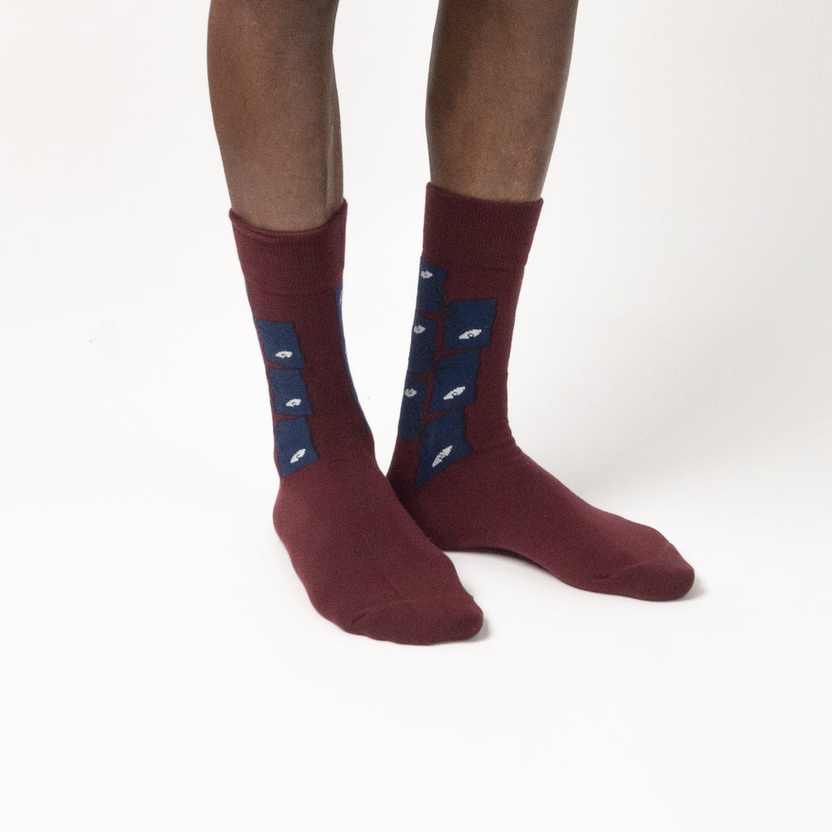 A pair of crew socks worn on a person's feet, shown against a plain white studio background. The socks are burgundy with a navy blue panel on the side featuring eye illustrations.