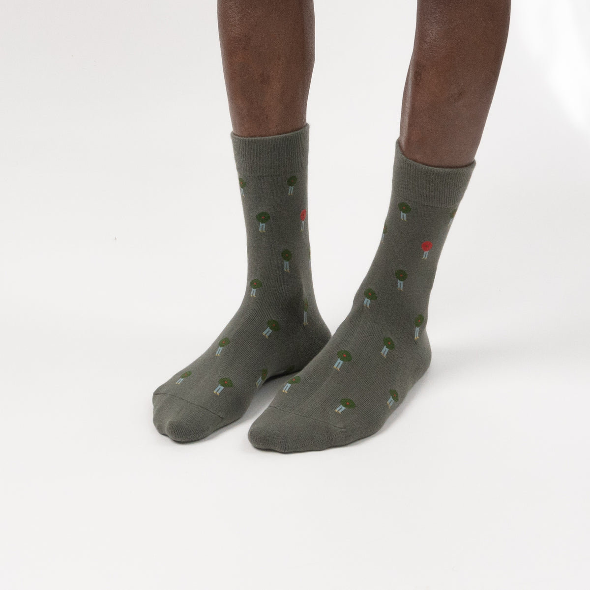 A pair of crew socks worn on a person's feet, shown against a clean white studio background. The socks are olive green with a small repeating pattern of tiny red, green, and white button illustrations.