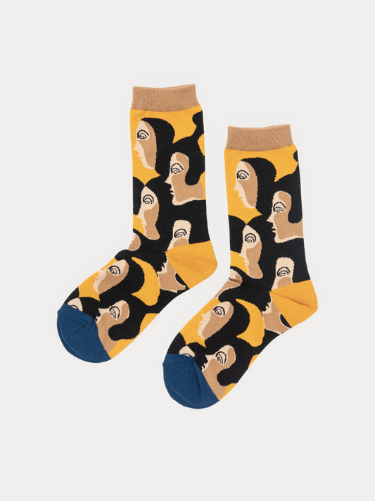 A pair of crew socks with a bold graphic pattern of stylized faces in black, beige, and yellow, featuring solid navy blue toes and heels, displayed on a light gray background.