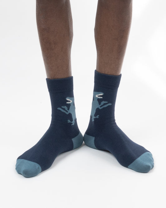 A pair of dark navy crew socks with a light blue dinosaur graphic and blue toe/heel accents, shown on a person's feet against a white background.