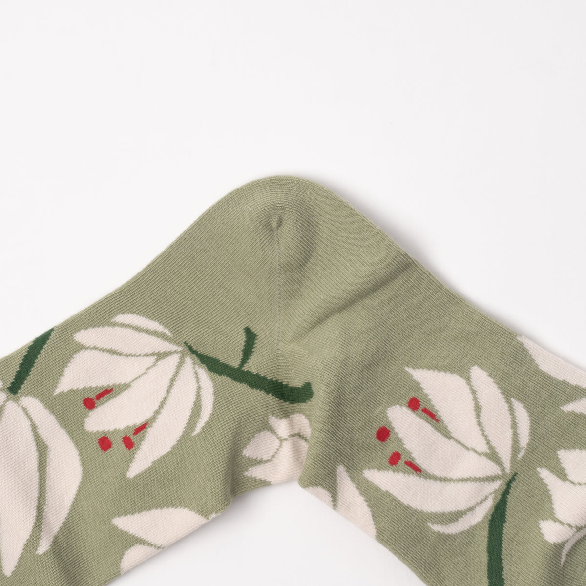 Close-up of a knit sock or cuff in muted sage green with a large white floral pattern and small red accents, photographed on a clean white background.
