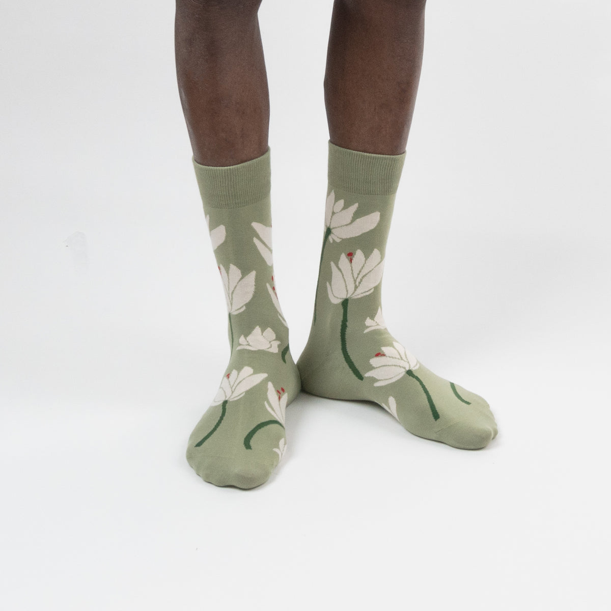A pair of sage green crew socks with a white lotus floral pattern, worn on a person's feet against a plain white studio background.