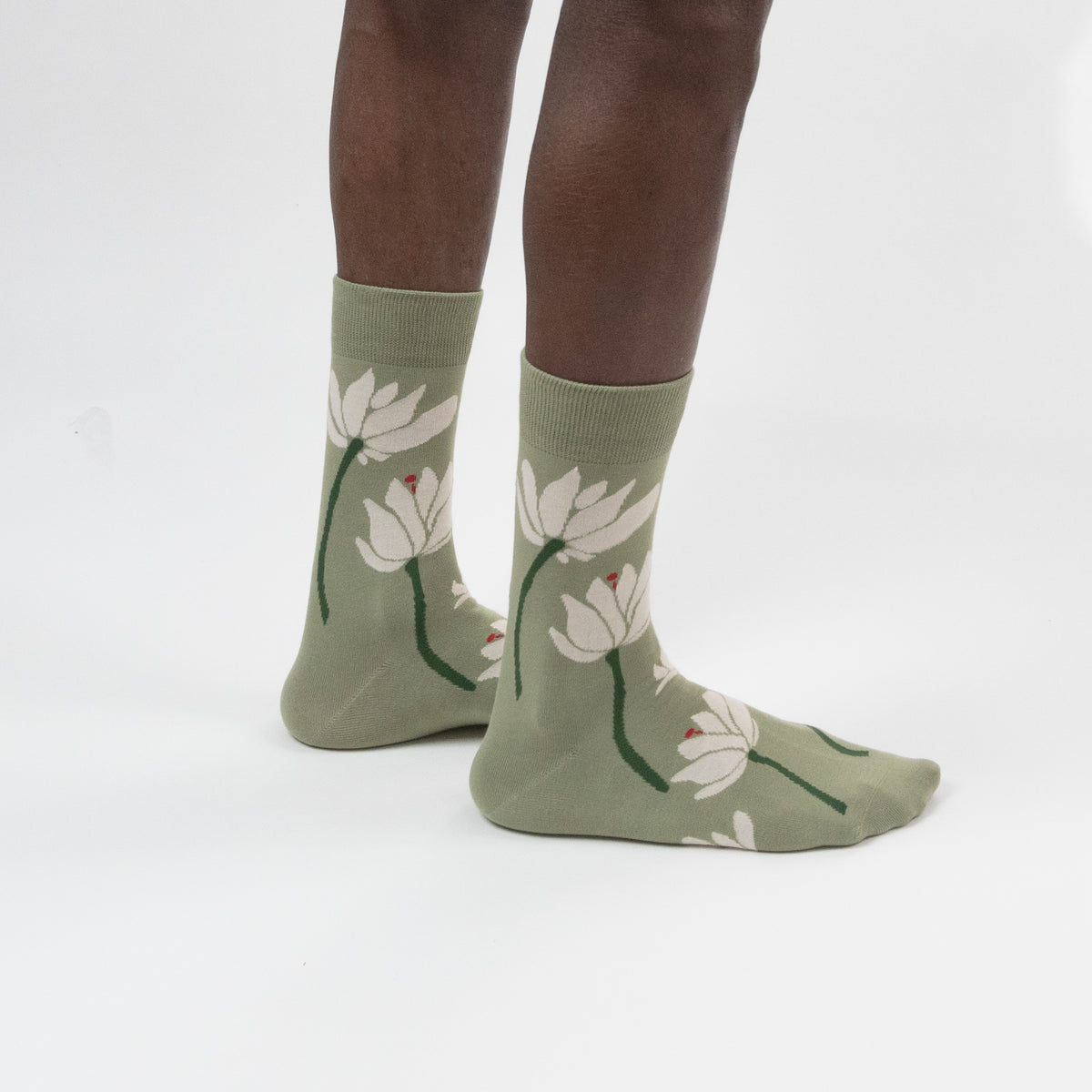 A pair of sage green crew socks with a white lotus floral pattern, worn on a person's feet against a plain white studio background.