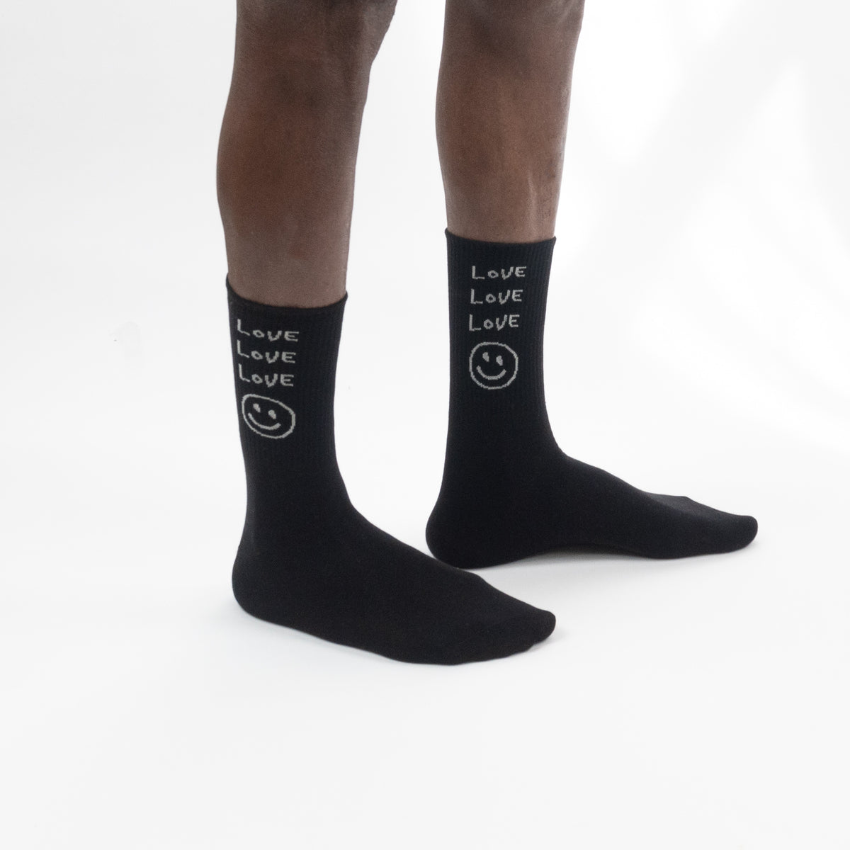 Black crew socks with white text reading 'LOVE LOVE LOVE' and a small smiley face, shown worn on a person's feet against a white studio background.
