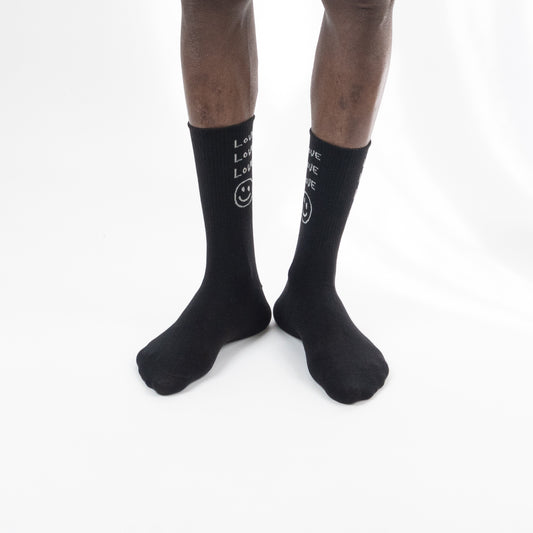 A person's legs wearing black crew socks with white text and a smiley face graphic, shown against a plain white background.