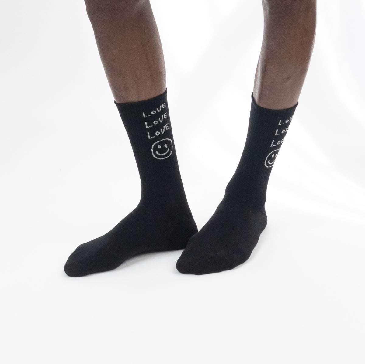 Black crew socks with white text reading 'Love Love Love' and a smiley face, worn on a person's feet against a plain white background.