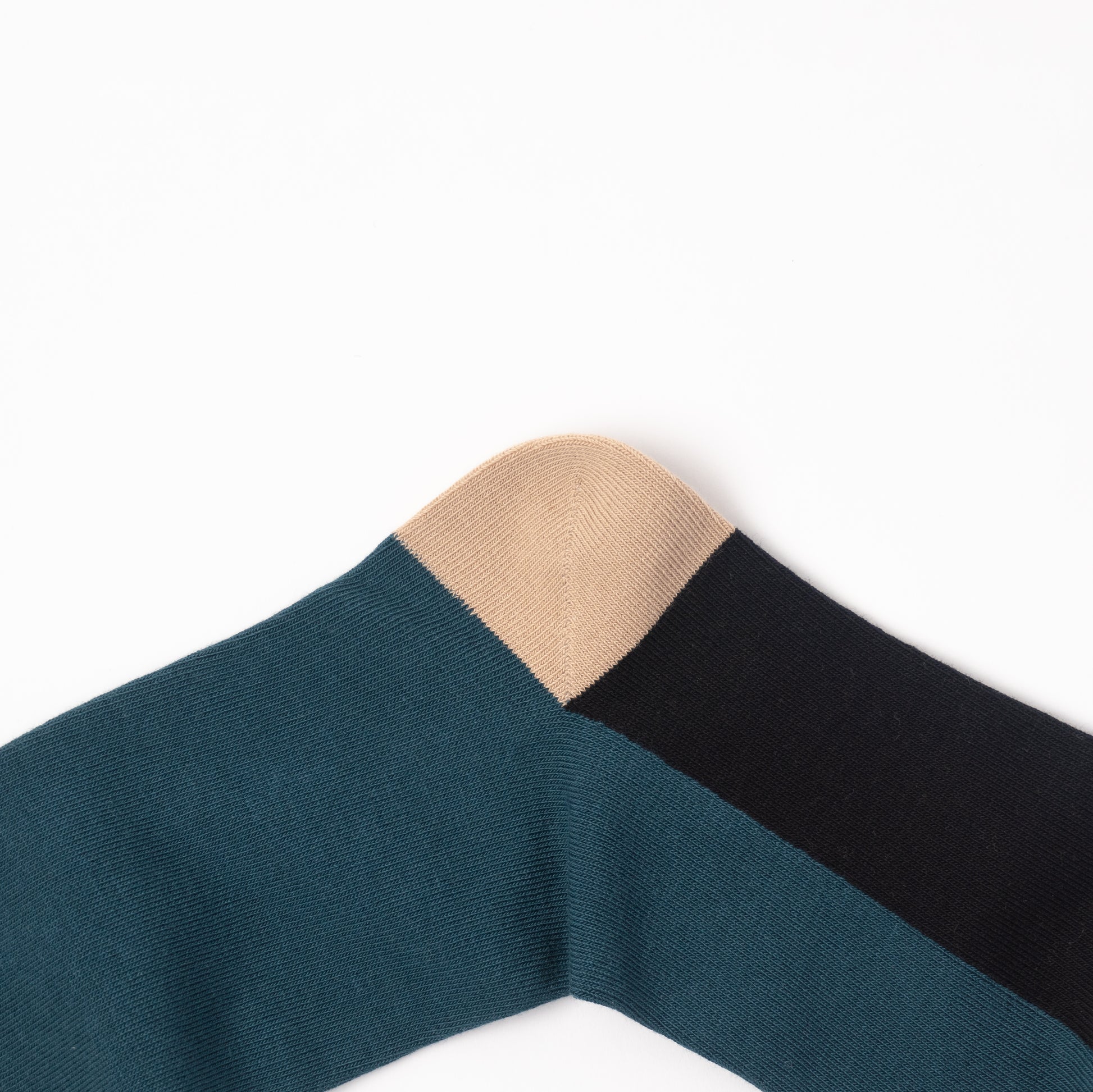 A close-up of a ribbed knit sock showing two color-blocked sections: a dark teal body and a black toe cap, with a beige heel section.