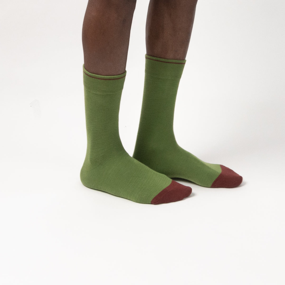 A pair of olive green crew socks with contrasting dark red toes, worn on a person's legs against a plain white studio background.