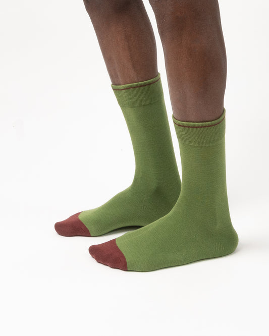 A pair of olive green crew socks with contrasting dark red toes, worn on a model's feet against a plain white background.