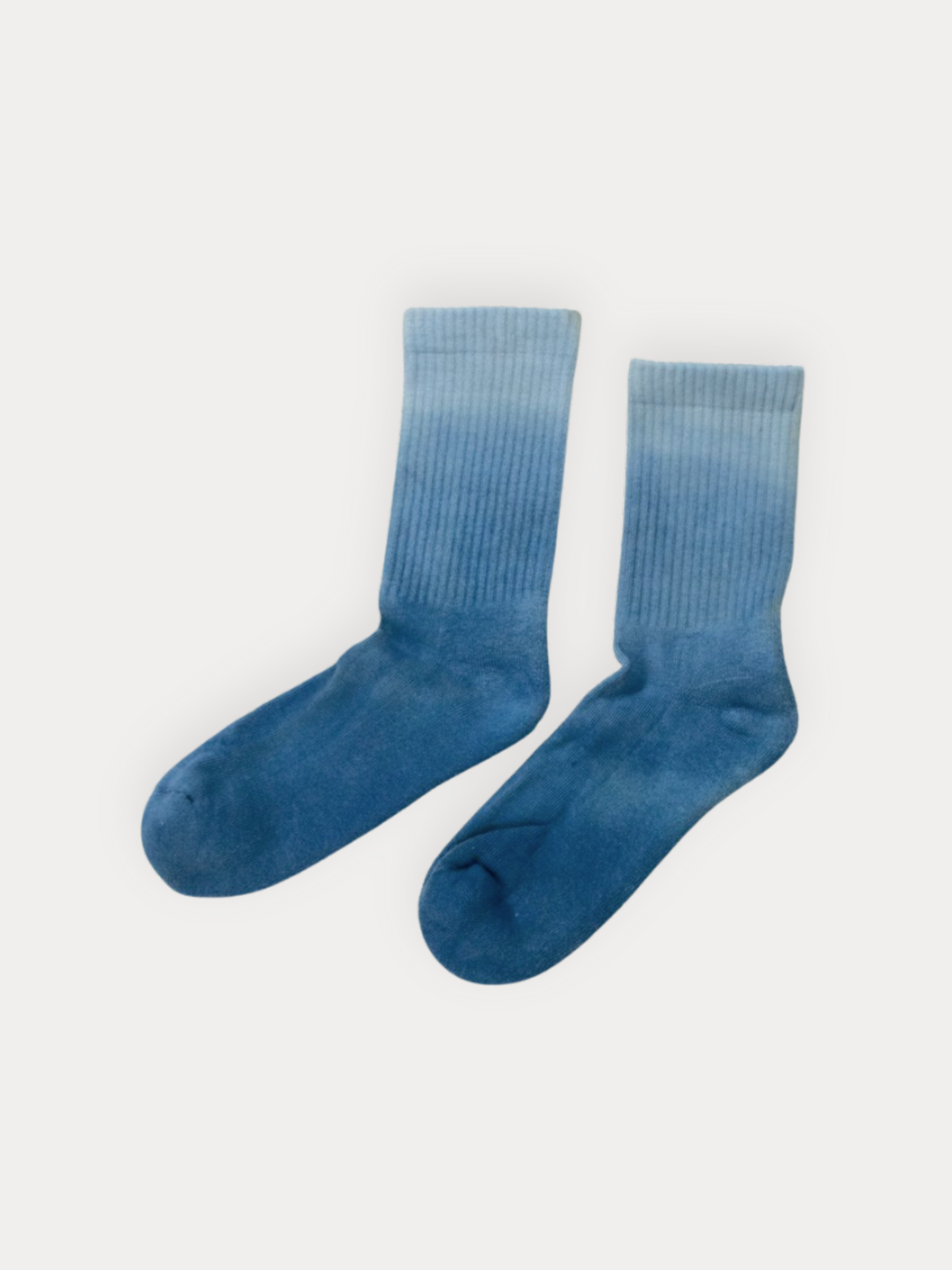 A pair of ribbed crew socks with a blue ombre gradient, fading from a lighter sky blue at the top to a darker medium blue at the bottom, shown on a light gray background.