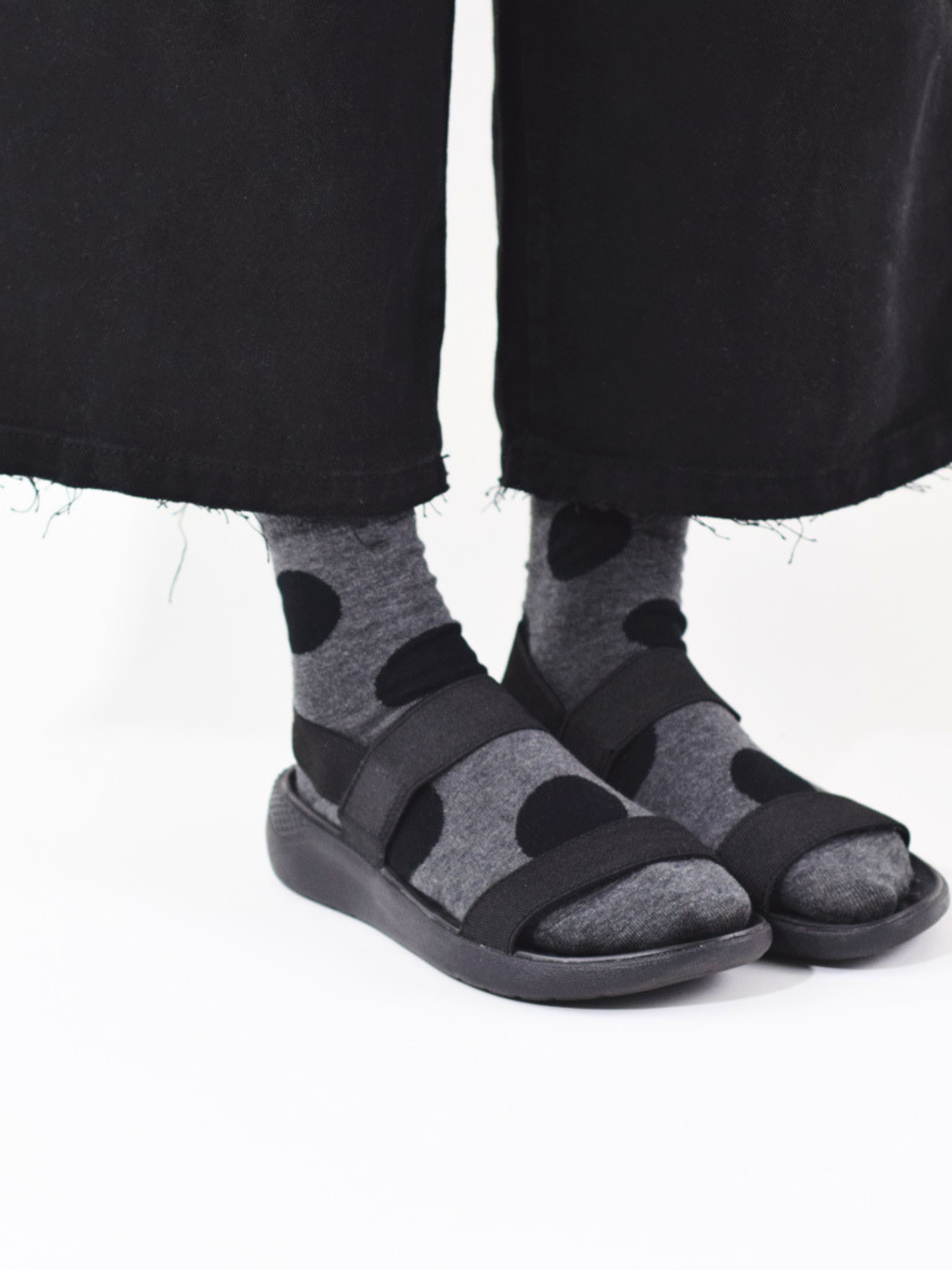 A close-up of a person's feet wearing dark gray and black polka-dot knit socks with black slide sandals, paired with black jeans against a white background.