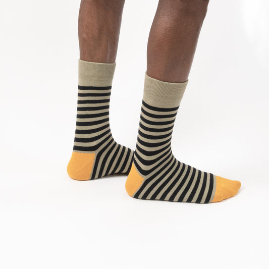 A pair of crew socks with black and beige horizontal stripes, featuring solid beige cuffs and mustard yellow toes and heels, shown on a model's feet against a white background.