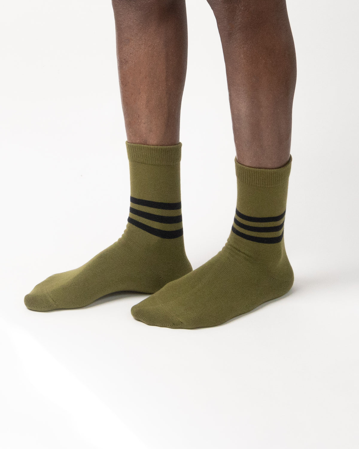 A pair of olive green crew socks with three black stripes near the cuff, worn on a person's feet against a clean white background.