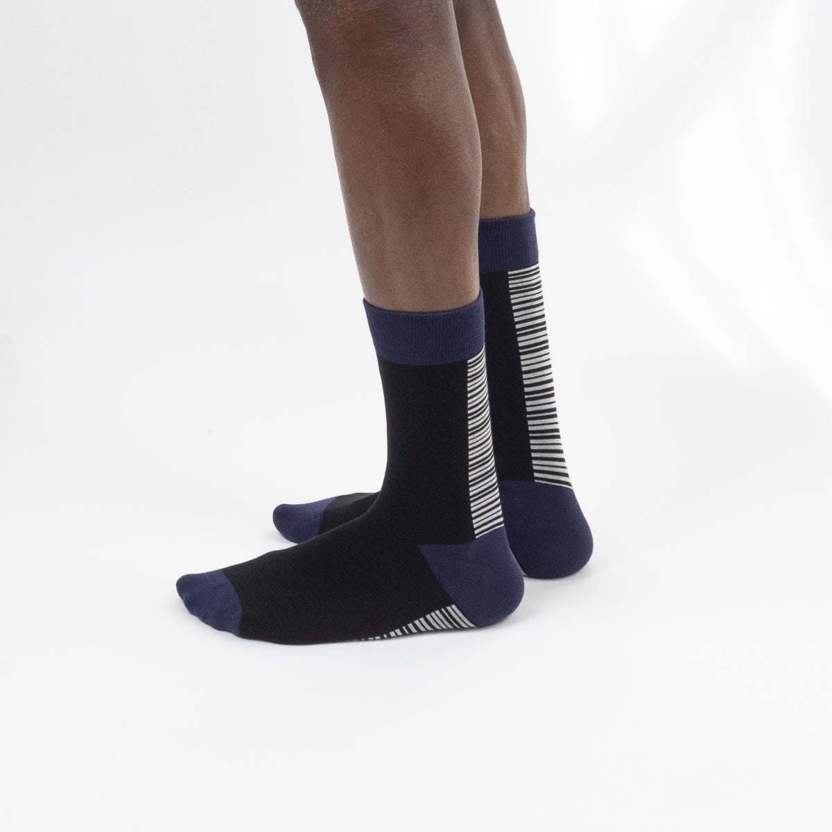 A pair of crew socks worn on a person's feet, shown against a clean white studio background. The socks are primarily black with navy blue cuffs, heels, and toes, and feature white vertical stripe patterns on the sides and soles.