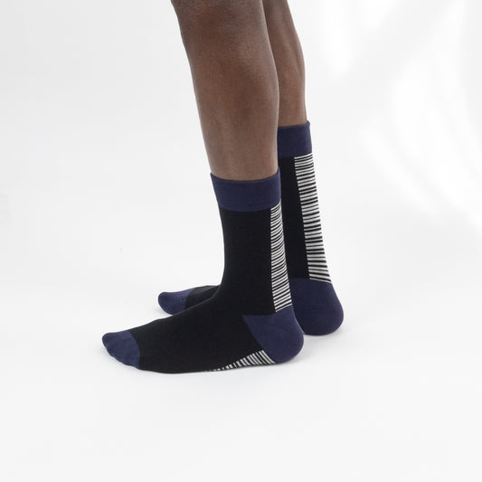 A pair of crew socks worn on a person's feet, shown against a clean white studio background. The socks are primarily black with navy blue cuffs, heels, and toes, and feature white vertical stripe patterns on the sides and soles.