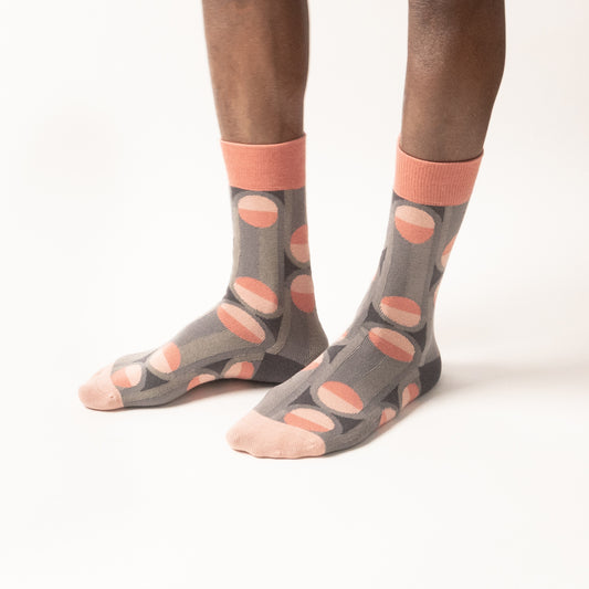 Model standing in grey crew socks with a two-tone coral and blush dot pattern, coral cuffs, and blush toes, against a white background.