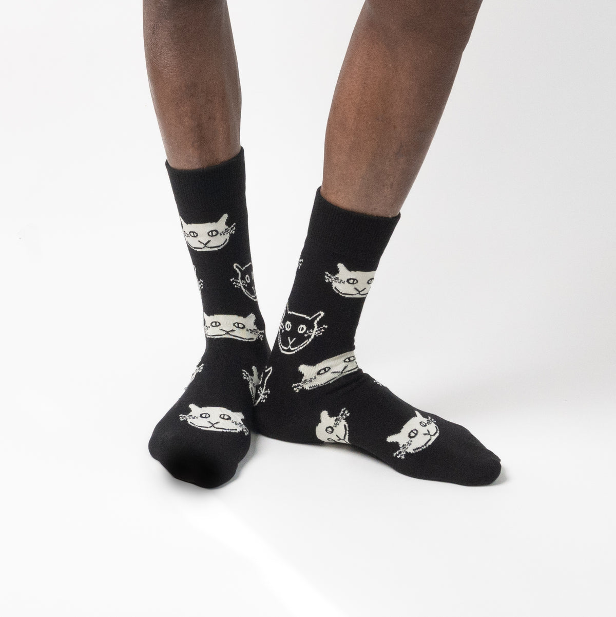 Black crew socks with a white cartoon cat face pattern, worn on a model's feet against a clean white studio background.