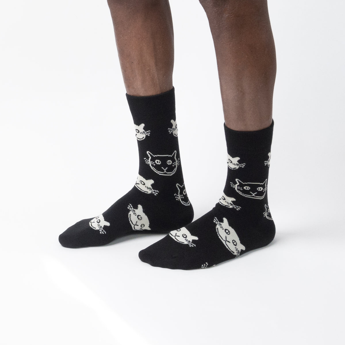 Black crew socks with a white cartoon cat pattern, worn on a person's feet against a clean white background.