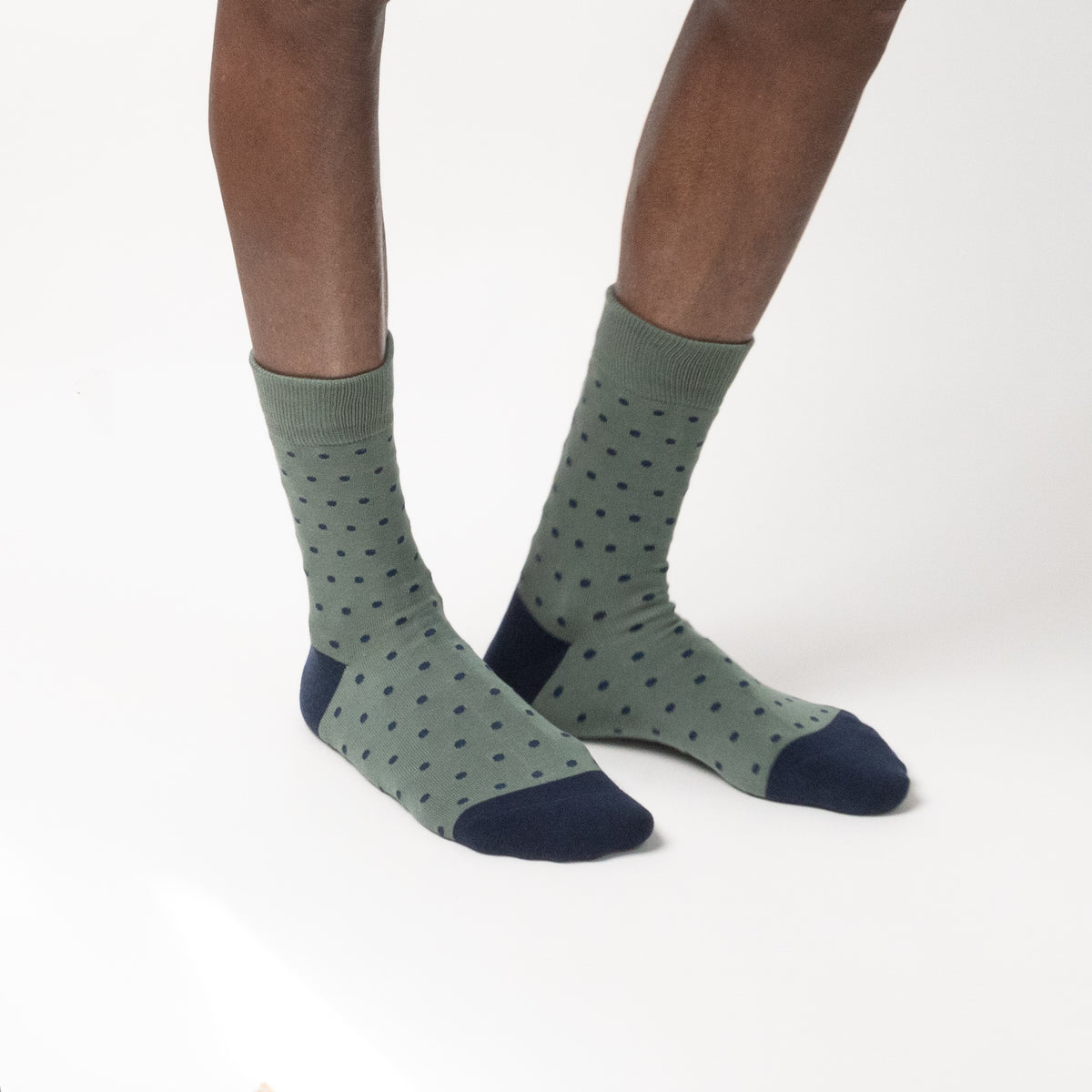 A pair of crew socks worn on a model's feet, featuring a sage green base with a pattern of small dark green dots and solid navy blue heels, toes, and cuffs, shown against a plain white studio background.