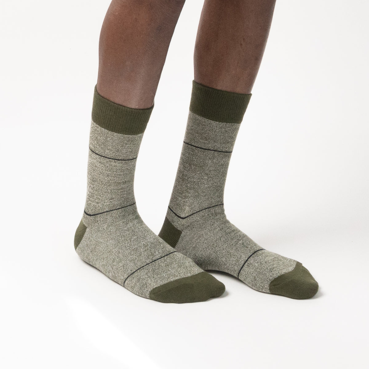 Three-quarter side view of a model wearing heather gray crew socks with thin horizontal black grid lines, featuring dark olive green cuffs, heels, and toes against a white background.