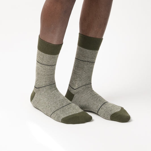 Three-quarter side view of a model wearing heather gray crew socks with thin horizontal black grid lines, featuring dark olive green cuffs, heels, and toes against a white background.