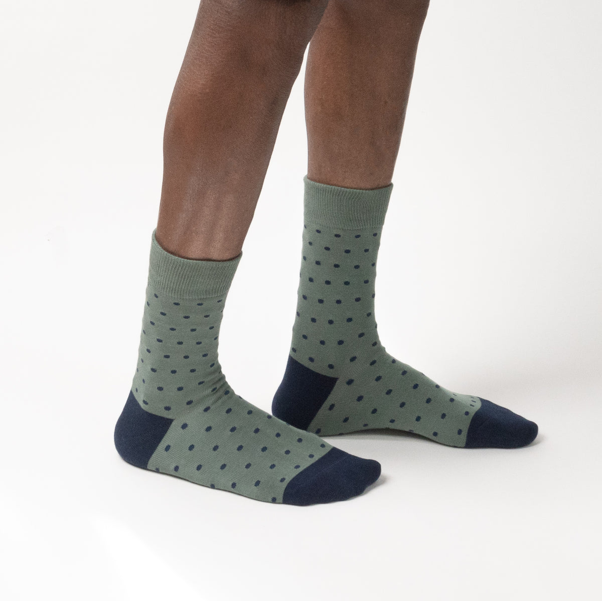 A model wearing a pair of crew socks in sage green with a dark green polka dot pattern, navy blue heels, and navy blue toes, shown against a plain white studio background.