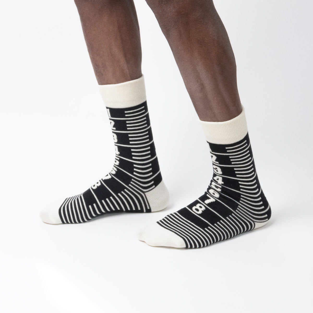 Three-quarter side view of a model wearing black and cream crew socks featuring a graphic ruler design with numbered markings from one to eight, standing on a white background.