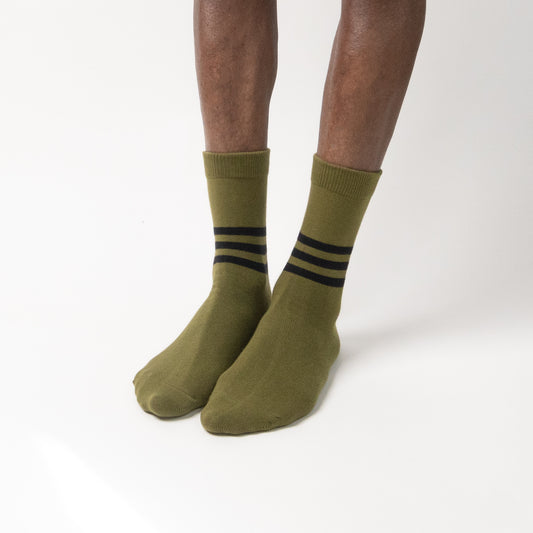 Front-facing view of a model wearing mid-calf olive green crew socks with three horizontal black stripes around the ankle, standing on a white background.